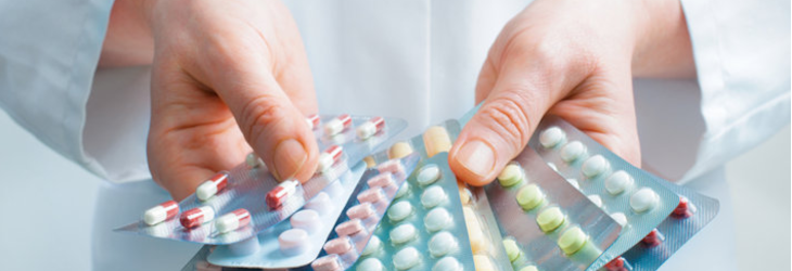 Some hands holding a variety of pills in different coloured blister packets. The person who owns the hands is wearing a lab coat and stethoscope